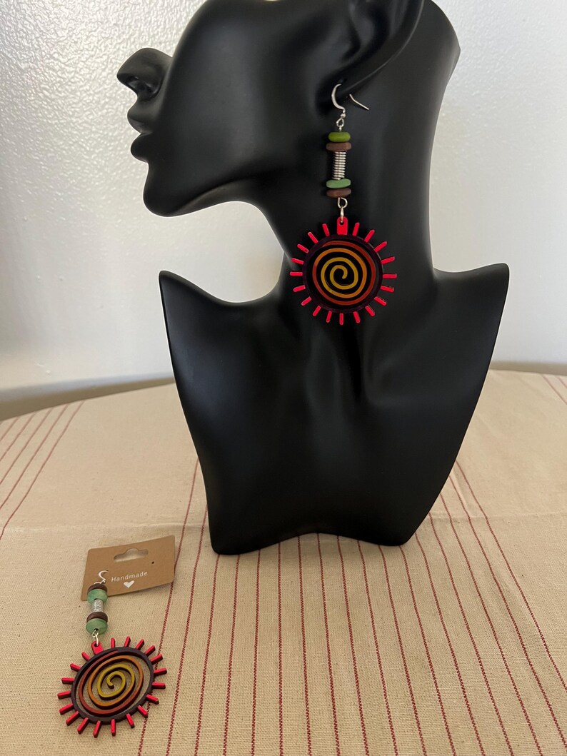May include: A pair of handmade wooden earrings with a red, orange, and yellow sun design. The earrings are hanging from a silver hook and have a green, brown, and silver bead dangle.
