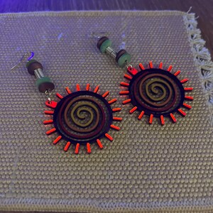 May include: A pair of earrings with a red, orange, and brown sun design. The earrings have a silver hook and a beaded dangle.