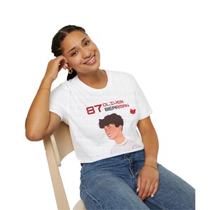 May include: A white t-shirt featuring a red and black graphic of a person with the text "87 Oliver Bearman" on it.