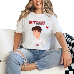 May include: White t-shirt featuring a graphic of a young man with the text "87 Oliver Bearman" printed on it. The graphic also includes a small red bear icon.