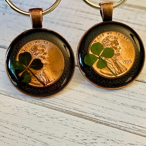 May include: Two round copper-colored keychains. Each keychain features a penny with a four-leaf clover and the text "IN GOD WE TRUST" and the year 1992 or 1995. The pendants have a black border and are attached to a silver key ring.