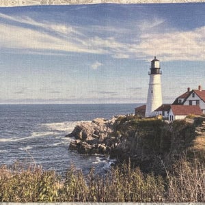 May include: A white lighthouse stands tall on a rocky cliff overlooking a blue ocean. A small sailboat is visible in the distance. The sky is a light blue with white clouds.