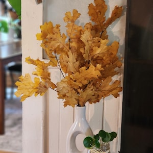 Könnte beinhalten: Eine weiße Keramikvase hält einen Strauß getrockneter gelber Eichenblätter. Eine kleine goldene Kerze steht auf einem weißen Teller, und ein kleines Glas mit grünen Pflanzen befindet sich neben der Vase. Die Anordnung steht vor einer weißen Wand.