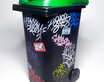 Pode incluir: Um pequeno caixote do lixo preto com tampa verde e base preta. O caixote está coberto de grafites coloridos em branco, rosa, azul, amarelo e vermelho. Uma pequena roda preta é visível.