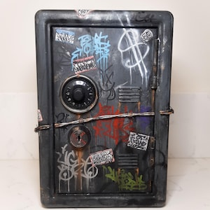 May include: A black metal safe with a combination lock and a keyhole. The safe is covered in graffiti and stickers, including a dollar sign and the word "UNDERCOVER".
