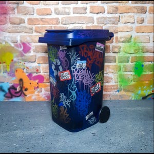 Op de afbeelding: Een donkerblauwe vuilnisbak met een deksel en een enkel wiel. De bak is bedekt met kleurrijke graffiti-kunst, waaronder tags en stickers. De achtergrond toont een bakstenen muur en meer graffiti.