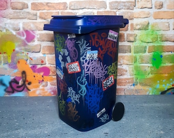 Pode incluir: Um caixote do lixo azul escuro com tampa e uma única roda. O caixote está coberto de arte de grafite colorida, incluindo tags e autocolantes. O fundo apresenta uma parede de tijolo e mais grafite.
