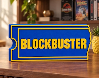 May include: A rectangular blue sign with a yellow border and a cutout on the left side, featuring the word BLOCKBUSTER in bold yellow capital letters. The sign has a raised, three-dimensional design and is positioned on a flat wooden surface.