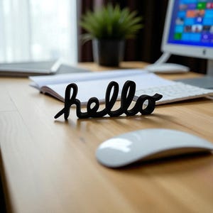 May include: A black cursive "hello" sign sits on a wooden desk, near a white computer mouse. A computer, keyboard, and a small potted plant are in the background. The sign is the focal point of the image.