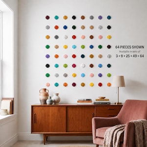 May include: A collection of 64 small, circular, flower-shaped wall decorations arranged in an eight-by-eight grid. Each piece features a distinct solid colour. Text on the right reads: 64 PIECES SHOWN Available in sets of 3 • 9 • 25 • 49 • 64.