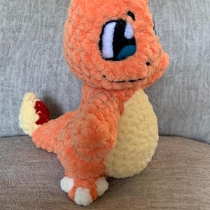 Op de afbeelding: Een oranje gehaakte Charmander-knuffel met een crème-kleurige buik en witte accenten op de voeten. De knuffel heeft grote blauwe ogen en een rood-gele vlam op de staart. Het speelgoed is gemaakt van zacht, getextureerd garen.
