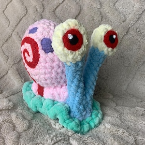 May include: A crocheted snail plush toy featuring a light blue body, a mint green base, and a pink shell. The shell is decorated with a red spiral and purple spots. Two tall stalks extend upward, each topped with a cream-colored eye with a red iris.