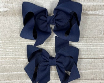 May include: Two navy blue hair bows, one larger than the other, on a light wood background.