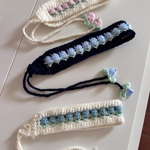 May include: Three handmade crochet headbands in cream, navy blue, and cream colors. Each headband features a floral design in pink, blue, and green. The headbands have long ties and are displayed on a white surface.