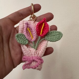 May include: A handmade crochet keychain featuring a bouquet of flowers. The bouquet includes pink and red calla lilies with yellow centers, green leaves, and a pink bow. The keychain has a gold-colored clasp.