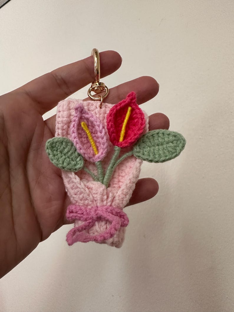 May include: A handmade crochet keychain featuring a bouquet of flowers. The bouquet includes pink and red calla lilies with yellow centers, green leaves, and a pink bow. The keychain has a gold-colored clasp.