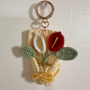 May include: A handmade crochet keychain featuring a small bouquet of flowers. The flowers include a white calla lily with a yellow center, a red flower, and green leaves. The keychain is attached to a gold-colored metal ring.