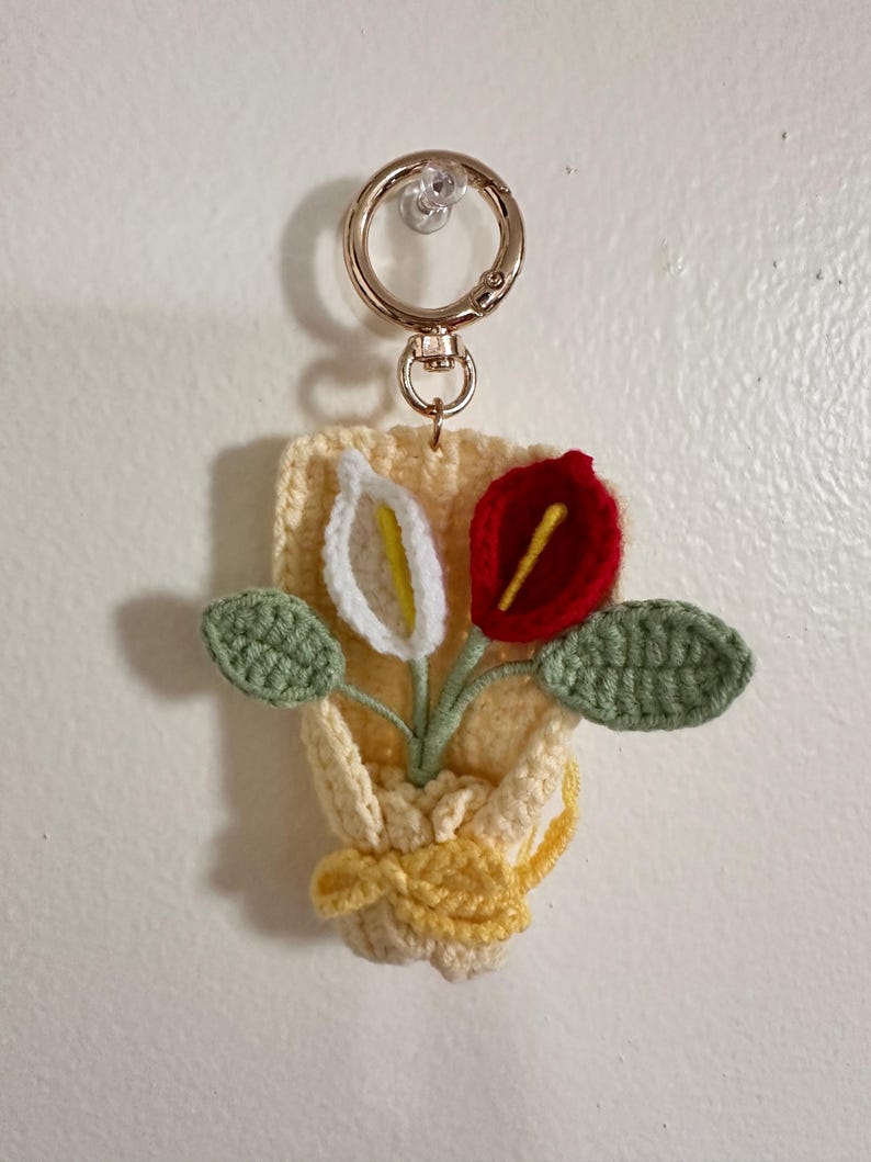 May include: A handmade crochet keychain featuring a small bouquet of flowers. The flowers include a white calla lily with a yellow center, a red flower, and green leaves. The keychain is attached to a gold-colored metal ring.