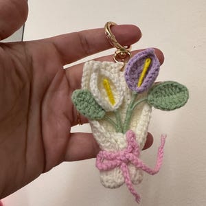 May include: A handmade crochet keychain featuring a bouquet of calla lilies. The flowers are white and purple with yellow centers, green leaves, and a pink bow. The keychain has a gold-colored clasp.