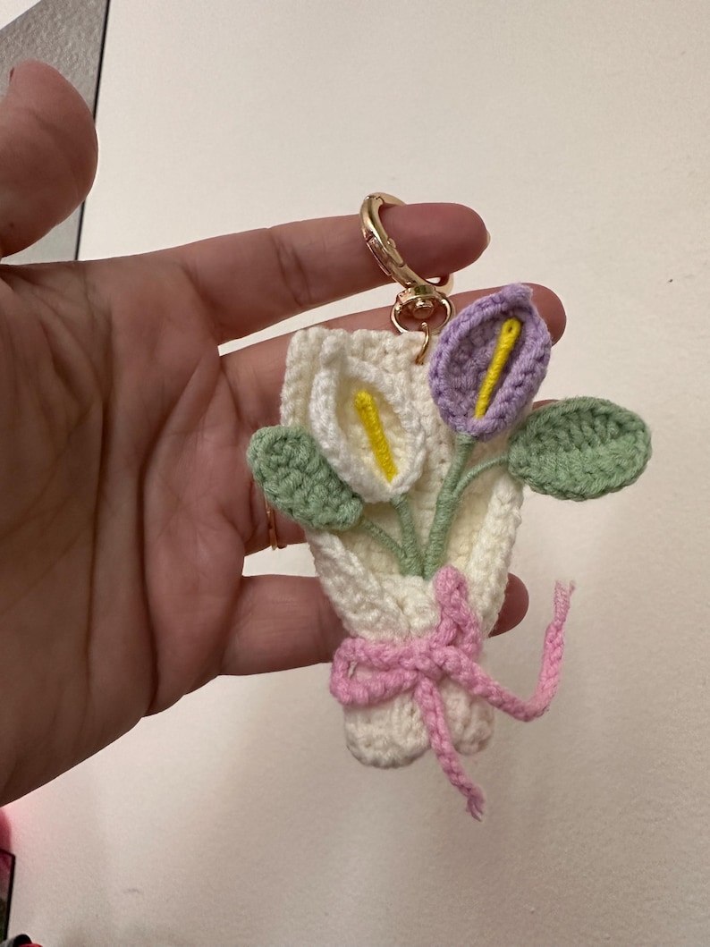 May include: A handmade crochet keychain featuring a bouquet of calla lilies. The flowers are white and purple with yellow centers, green leaves, and a pink bow. The keychain has a gold-colored clasp.