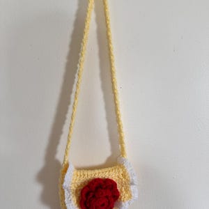 May include: A handmade, yellow crochet purse with a long strap. The purse features a red flower in the center and white ruffled trim. The bag is hanging against a white wall.