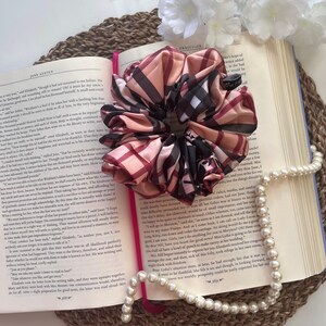 Könnte beinhalten: Ein rosa-rotes Karoscrubbie mit einem schwarz-weißen Karomuster. Das Scrunchie liegt auf einem Buch mit dem sichtbaren Text "Pride and Prejudice". Eine weiße Perlenkette liegt neben dem Buch auf einer geflochtenen Oberfläche.