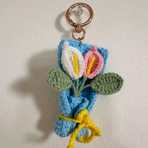 May include: Crocheted flower bouquet keychain featuring white and pink flowers with yellow centers and green leaves. The bouquet is wrapped in blue yarn and tied with a yellow ribbon. A gold-colored keyring is attached.