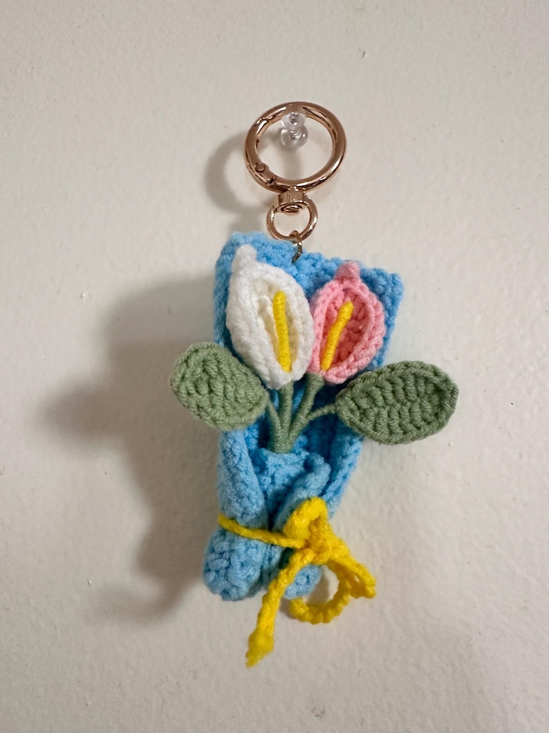 May include: Crocheted flower bouquet keychain featuring white and pink flowers with yellow centers and green leaves. The bouquet is wrapped in blue yarn and tied with a yellow ribbon. A gold-colored keyring is attached.