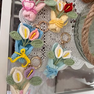 May include: A collection of handmade crochet flower bouquet keychains in various colors. Each keychain features a small bouquet of flowers with green leaves, tied with a ribbon or bow, and attached to a gold-colored keyring. The bouquets are in shades of pink, yellow, blue, and white.