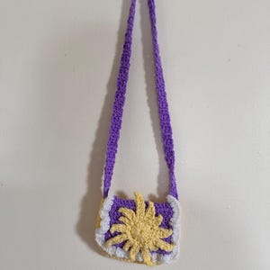 May include: A handmade crochet purse with a long strap, in purple, white, and yellow. The purse features a sun design on the front, with a purple body, white trim, and a yellow sunburst. The strap is also purple.