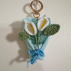 May include: A handmade crochet keychain featuring a bouquet of calla lilies. The bouquet is in a light blue holder with a blue bow. The flowers are white with yellow centers and green leaves. A gold-colored keyring is attached.