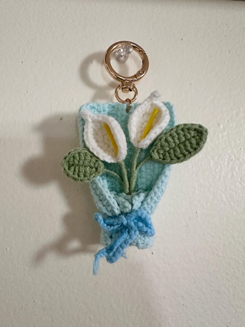 May include: A handmade crochet keychain featuring a bouquet of calla lilies. The bouquet is in a light blue holder with a blue bow. The flowers are white with yellow centers and green leaves. A gold-colored keyring is attached.