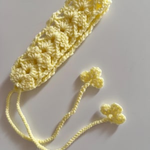 May include: A handmade, pale yellow crochet bookmark with a floral design. The bookmark has two dangling flower accents. The crochet work is visible, showcasing the texture and craftsmanship of the item. The bookmark is lying on a white surface.