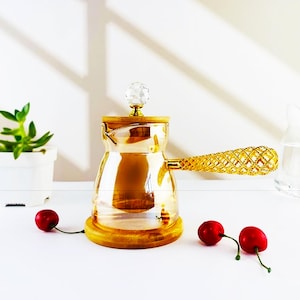 May include: A clear glass coffee pot with a gold-coloured handle and base. The handle has a textured design. The lid is topped with a crystal-like knob. Two red cherries are in the foreground. A small potted plant is in the background.