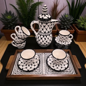May include: A black and white ceramic coffee set with a geometric pattern. The set includes a tall coffee pot, several small cups, saucers, and a serving tray. The cups and saucers are stacked and arranged on a wooden tray.