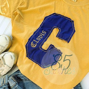 May include: Mustard yellow t-shirt with a large blue letter "C" patch. The patch has gold stitching and the name "Clovis" embroidered on it. The shirt is paired with blue jeans and white sneakers.