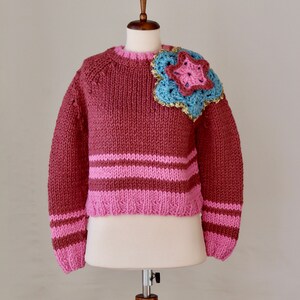 May include: A cropped, red sweater with pink stripes and a crocheted flower applique on the shoulder.