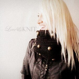 May include: A woman with long blonde hair is wearing a brown knit cowl neck with gold buttons. The cowl neck is a chunky knit and has a textured look.