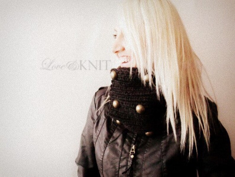 May include: A woman with long blonde hair is wearing a brown knit cowl neck with gold buttons. The cowl neck is a chunky knit and has a textured look.