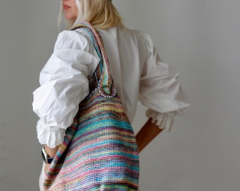 Könnte beinhalten: Eine Frau trägt ein weißes Hemd und eine helle Jeans und trägt eine große, bunte, gestreifte Crochet-Tragetasche. Sie trägt eine blaue Jeansmütze.