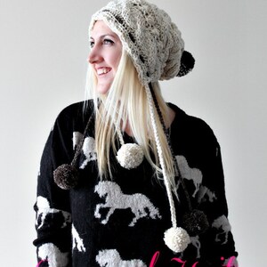 Può includere: Un berretto bianco lavorato a maglia con un pon pon in cima e lunghi lacci. Il berretto ha un motivo a treccia e viene indossato da una persona con lunghi capelli biondi. La persona indossa un maglione nero con un motivo a cavallo bianco.