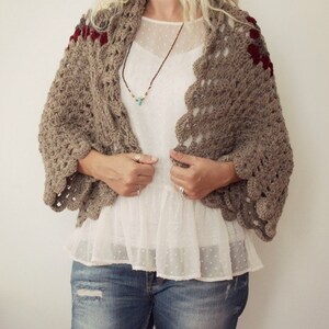 May include: A brown crocheted shawl with red and gray accents. The shawl has a scalloped edge and is worn over a white blouse and blue jeans.