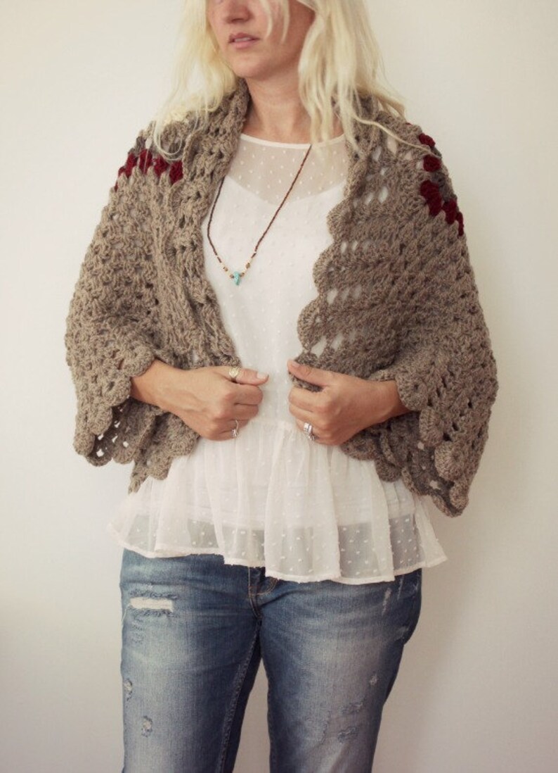 May include: A brown crocheted shawl with red and gray accents. The shawl has a scalloped edge and is worn over a white blouse and blue jeans.