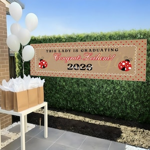 May include: A graduation banner with the text "THIS LADY IS GRADUATING Congrats Leilani! 2026" in red and black. The banner has a ladybug design and is decorated with red polka dots. White balloons and gift bags are also visible.