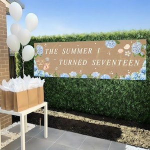 Könnte beinhalten: Ein Banner mit dem Text "THE SUMMER I TURNED SEVENTEEN" ist mit blauen Hortensien, Muscheln und Seesternen verziert. Weiße Luftballons und Geschenktüten sind in der Nähe angeordnet und schaffen eine festliche Atmosphäre. Das Banner wird vor einer grünen Hecke präsentiert.