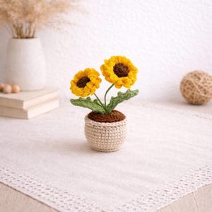 May include: A crocheted sunflower arrangement in a small, beige pot. The sunflowers have bright yellow petals and brown centers, with green leaves. The pot sits on a white lace-trimmed surface, with a vase and books in the background.