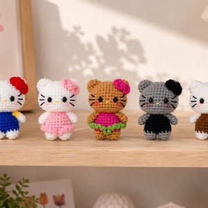 May include: Five small crocheted cat figures arranged in a row. The first is white with a red bow and blue dress, the second is white with a pink bow and dress, the third is brown with a pink bow and green skirt, the fourth is gray with a black bow and dress, and the fifth is white with a brown bow and dress.