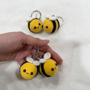May include: Handmade crochet bee keychains. Each bee is yellow with black stripes and white wings. The bees have smiling faces and are attached to silver keyrings. The keychains are a cute and whimsical accessory.