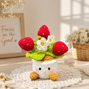 May include: A crocheted strawberry plant in a white pot with a smiling face. The plant features three red strawberries with white embroidered seeds, green leaves, and white flowers with yellow centers. The pot has two small brown feet at the base.