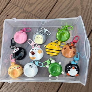 May include: A collection of eleven colorful, circular character-themed keychains featuring various designs including a pink pig, a grey creature, a green frog, a black figure, a white cat, a yellow bee, a brown bear, a tan rabbit, a white seal, a green turtle, and a black penguin.